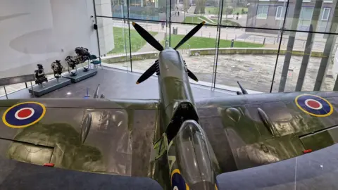 Stoke-on-Trent City Council A Spitfire plane, viewed from above, is in a large glass-fronted building that also contains mechanical parts of the aircraft on display.