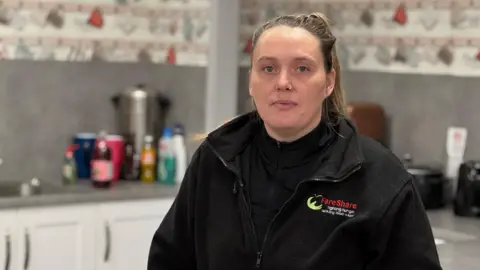 Naomi Winter with brown hair tied back in a ponytail, with a nose ring, wearing a black fleece that has the words 'fareshare' on the chest, stands in a kitchen. 