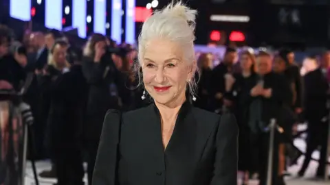 A woman with white hair smiles. She is wearing a black blazer, and behind her are photographers. 