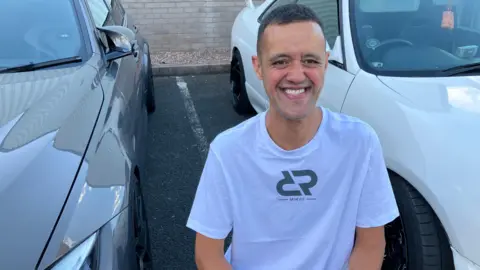 Reece is kneeling on the ground between two cars and is smiling into the camera. He wears a white T-shirt with a small black design on it and has short black hair.