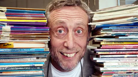 A grinning Michael Rosen with his head between two tall piles of books.