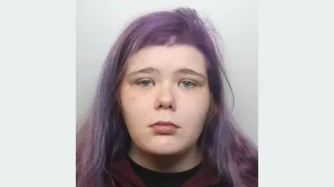 Avon and Somerset Police A police mugshot of Katherine Reilly, wearing a black t-shirt underneath a dark maroon hoodie. She has light blue eyes and long straight hair which is dyed purple. She is looking at the camera with a forlorn expression.