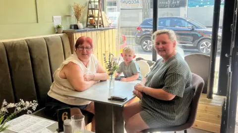 Two women sit in a cafe, facing each other, smile for the camera. Sitting in a highchair is a young boy with blond hair.