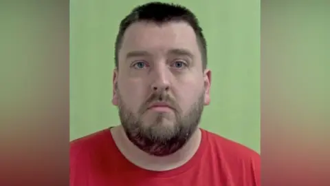 Police mugshot of Liam Bradley, who has short brown hair and a beard and is wearing a red T-shirt. He is set against a green background.