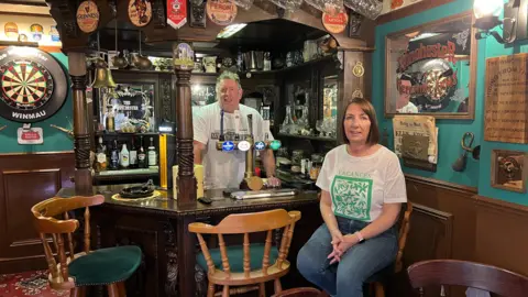 BBC Kev Marchant standing behind his small bar of a little pub room with Gina Marchant sitting on a barstool in front of it. The walls are painted green and adorned with an assortment of items including beer mats, a dart board and horse brasses.