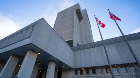 An image of the Ontario Court of Justice in London, Ontario. The photo is taken from a low angle, showing the concrete building which is grey concrete with square columns and a small tower in the centre. On the side of the building are two flag poles, one with a Canadian flag and one with the flag of Ontario. 