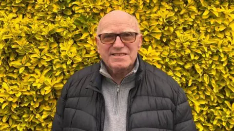 Rhys Thomas/BBC Mike Higgins is wearing a black padded jacket and grey zip up top in front of a hedge with yellow leaves.