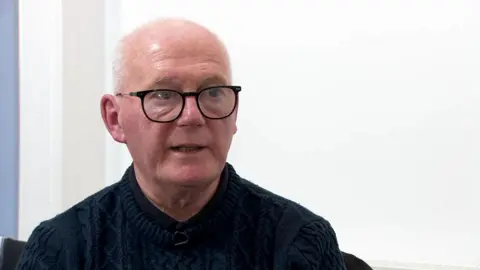 Paul O'Neill is bald with grey hair at the sides and is wearing black-framed glasses and a dark jumper and shirt 