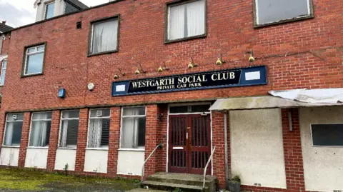 Exterior of Westgarth Social Club. It is a two-storey red bricked building with additional doormer windows in the roof. The windows on the ground floor are rectangular while the first floor ones are square. A long rectangular black sign above the central double doors gives the name of the venue and advertises a "private car park".