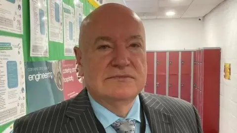 A bald man wearing a striped grey suit, blue shirt and silver tie. he is stood in a school corridor with a notice board on one side and red lockers on the other.