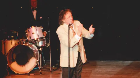 Embassy Theatre Ken Dodd, a man with a mop of brown hair wearing a cream jacket, bright patterned tie and black trousers, gestures with his hands as he stands on stage behind a microphone. Behind him, a man wearing a dinner jacket and bow tie sits behind a drum kit in front of a black backdrop.