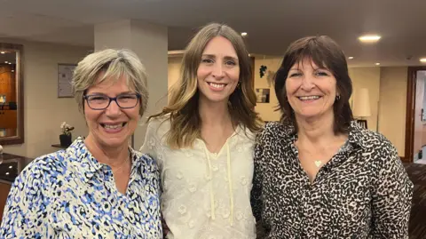 Harriet Heywood/BBC Three women are standing side by side indoors, smiling warmly at the camera. On the left is Ms Burgess wearing glasses and a blue and white patterned shirt. In the centre is Ms Harris with long hair, dressed in a white blouse with floral detailing. On the right is Ms Brookes, wearing a black and white patterned top and a heart-shaped necklace - that was gifted to her by Ms Harris. The setting is a bright, welcoming room with soft lighting and some furniture and decorations in the background.
