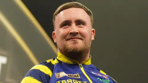 PA Media Luke Littler, pictured in March 2026, wearing a blue and yellow jacket during a darts tournament