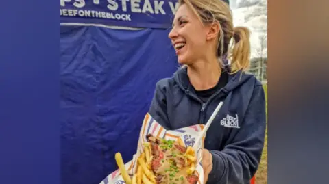 A woman holds a portion of steak and chips in a striped paper wrapper. She is wearing a blue zip-up top and standing in front of a blue stall outdoors.