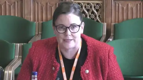 PA Media A brown haired woman wears black clear-lensed glasses. She wears a black t-shirt under a red cardigan. She has an orange lanyard around her neck. Behind her is a brown wood panelled wall with green seats in front of it.