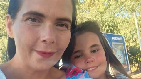 Emma Honeyfield Close up selfie of Emma and her daughter Amber smiling. Emma, on the left, has long dark hair and wears a blue and pink floral top. Amber has long light brown hair. 