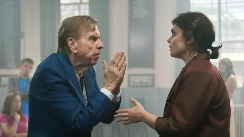 BBC/ BBC Studios John and Janey argue with one another in what appears to be a community hall. 