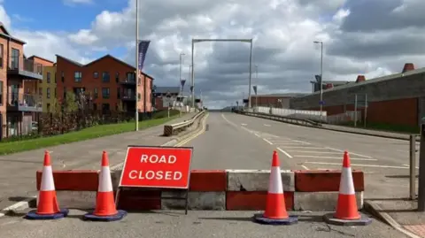 A road onto a bridge has a 'road closed' sign, with four orange cones and a little white and orange barrier. To the left of the road are blocks of flats.
