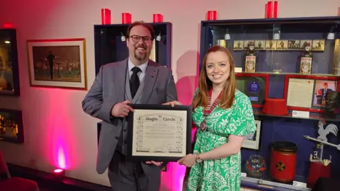 Peter Meacham Peter Meacham standing with a female friend, the two of them holding a framed Magic Circle certificate, with a display cabinet behind them