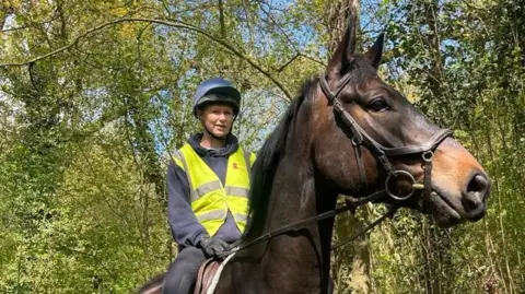 Amy Hilton A woman wearing a helmet and a high-visibility jacket riding a brown horse. There are green trees in the background. 