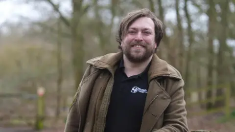 Jamie Niblock/BBC A man, Daniel Wade is standing in Fenland wearing a Suffolk Wildlife Trust t-shirt and army green coloured jacket. He has a beard and is smiling at the camera. 