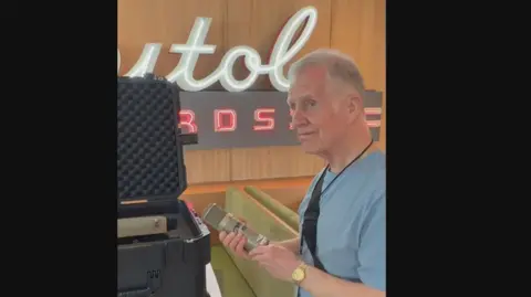 Jim Moir Jimmy looks emotional as he holds an old school chubby recording microphone beside its heavy flight case. The Capitol Records sign is behind him on the wall.