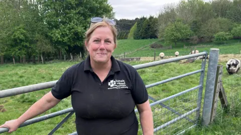 Kate Bradbrook/BBC Woman in black t-shirt smiles in front of five bar gate. The background is a field of sheep and pigs.