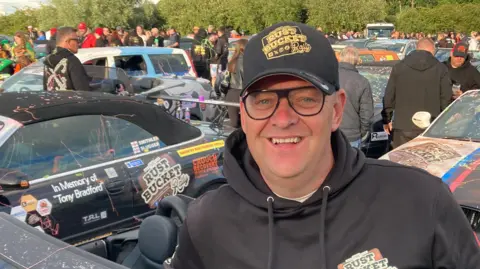 A man in a black hoody and black baseball cap surrounded by cars taking part in the challenge.