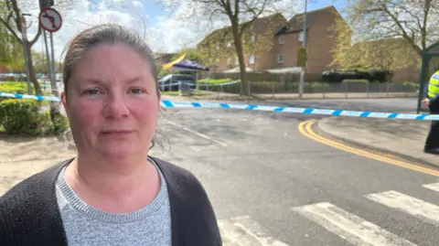 Kirsty Knight wearing a grey T-shirt and a black cardigan. She is standing in front of the crime scene. There is blue-and-white police tape in the background.