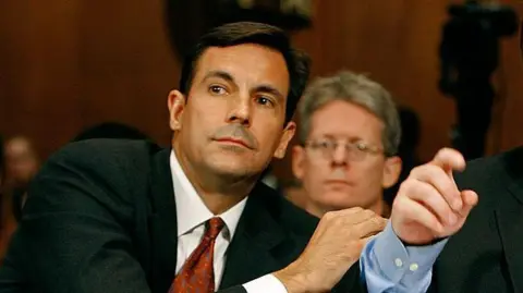 Getty Images Mark Paoletta, who has dark brown hair and wears a black suit with red tie, leans to the left to speak to his client during a Senate hearing in 2007