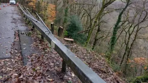 Devon County Council Shows part of the B3212 road on the left and a barrier that has been damaged with a steep hill going into woodland