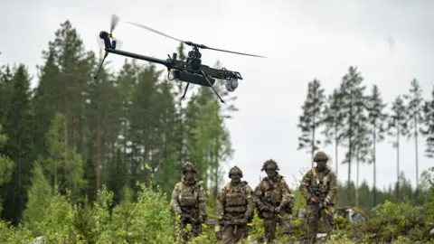Four soldiers watch an unmanned drone. They are standing on the edge of a wooded area with big trees.
