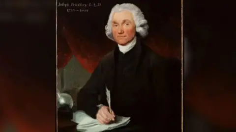 Dr Williams's Trust London/The Leeds Library A painting of Joseph Priestley holding a quill and writing on a piece of paper. He is wearing a black coat with a white collar and has white curly hair.