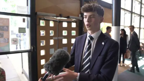 A young man with dark hair, wearing a blue suit and blue and white tie, speaks to a person holding a microphone