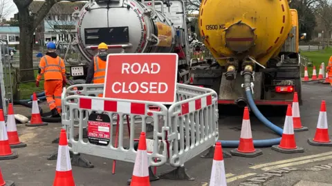 Eddie Mitchell A road closed sign and tankers pumping sewage