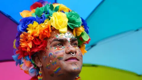 A person stands in front of a rainbow-coloured background. They have a rainbow flower headdress, multiple piercings and glitter and rainbow artwork on their face.