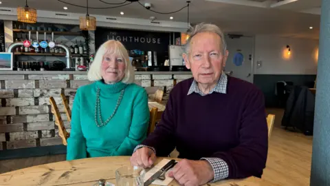 A woman in a green polo jumper sits next to a man wearing a plum jumper with a checked shirt. They are seated at a wooden table in The Lighthouse restaurant. 