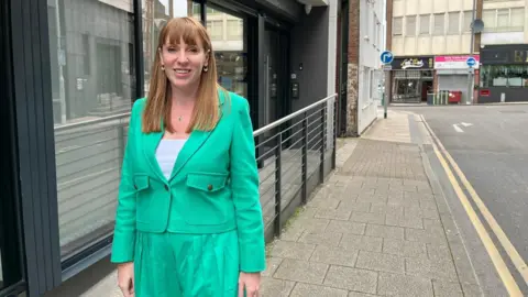 Angela Rayner in Hanley wearing a light green top and trousers over a white top while standing on pavement next to a building with a railing outside