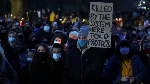 Reuters A woman at the gathering in Clapham Common hold a sign that reads: "Killed by the system we're told protects"