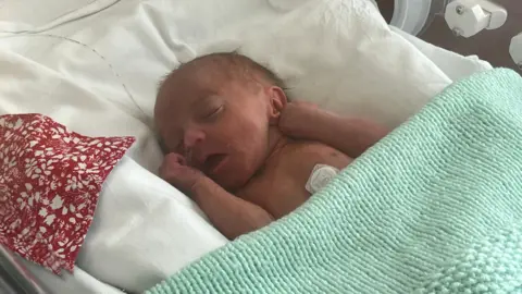 Baby Noah in a cot. He is sleeping, tucked under a mint-coloured blanket. His mouth is open and his hands are by his face. 