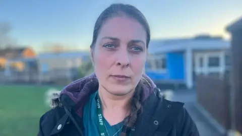 BBC / Elizabeth Baines Mum, Sibel Ansell looks at the camera. She is wearing a green top and black coat. Her brown hair is in a side plait. The school is in the background.