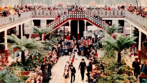 Glasgow City Council A group of dancers parade through the Springburn Winter Gardens as crowds watch on from above. Large plants are in either side of the aisle.