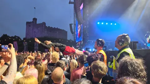 Dave Marlow Jaret Reddick son, Everett Reddick doing a crowd surf during the Bowling for Soup show at Cardiff Castle. Everett is pictured being heled up by fans, with his arms in the air. He is wearing a black hoodie and red shorts. Three team members (two men and one woman) can be seen standing by as he is lifted. In the far left of the image, Cardiff Castle can be seen in the background. The stage can be seen in the far right of the image.