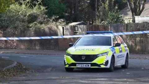 A police car and a police tape stretched across the road, Heol Trostre, in St Mellons, Cardiff. A house is in the background, and trees and fencing