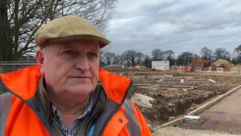 Andrew Turner/BBC Alistair Beales, wearing a tweed flat cap, orange high-visibility jacket and checked shirt. He is standing next to a construction site.