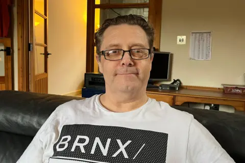 Charlotte Rose/BBC Mike Williams sitting in his living room in Caterham wearing a white tee shirt and spectacles.