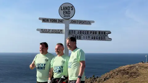 Licensed Trade Charity/Platinum Live Gary McKee standing at Land's End with two members of his running support team. All are wearing mint green T-shirts with the words "lucky saint" on the front. They are standing next to a black and white finger signpost detailing distances from Land's End to other places, such as New York and John O'Groats. The men are pictured side-on posing for a picture. They are all smiling and one is giving the camera a thumbs up.
