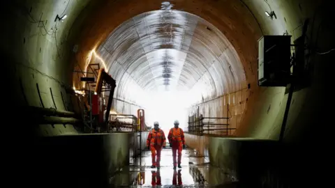 Two workers in orange safety gear walk through a large railway tunnel, with reflective water on the floor and bright daylight visible behind them.