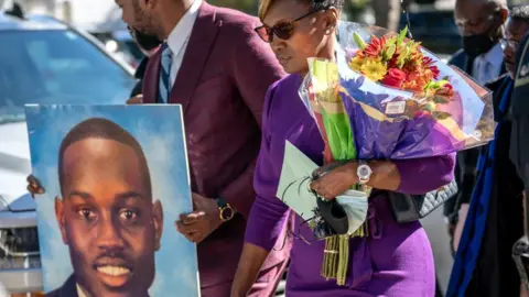 Wanda Cooper-Jones, wearing sunglasses and a purple dress, and holding a bouquet of flowers, walks out of the Glynn County Courthouse looking down, as a man to her right carries a portrait of Ahmaud Arbery