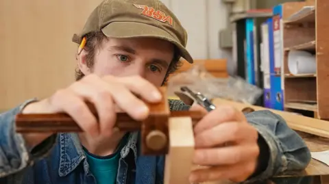 Martin Giles/BBC Hector is wearing a blue denim jacket, and black hat. His eyes peer onto his hands, where he is using a tool to make markings on his work. 